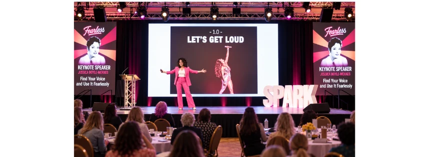 Jessica Doyle-Mekkes on stage in a pink suit, arms open, at the Spark Women's Leadership Conference.